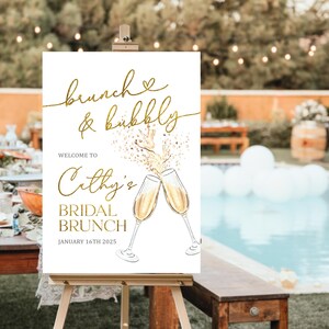 May include: A white sign with gold lettering and illustrations, displayed on a wooden easel. The sign reads "brunch & bubbly" and "Cathy's Bridal Brunch, January 16th 2025". Two champagne glasses are illustrated.