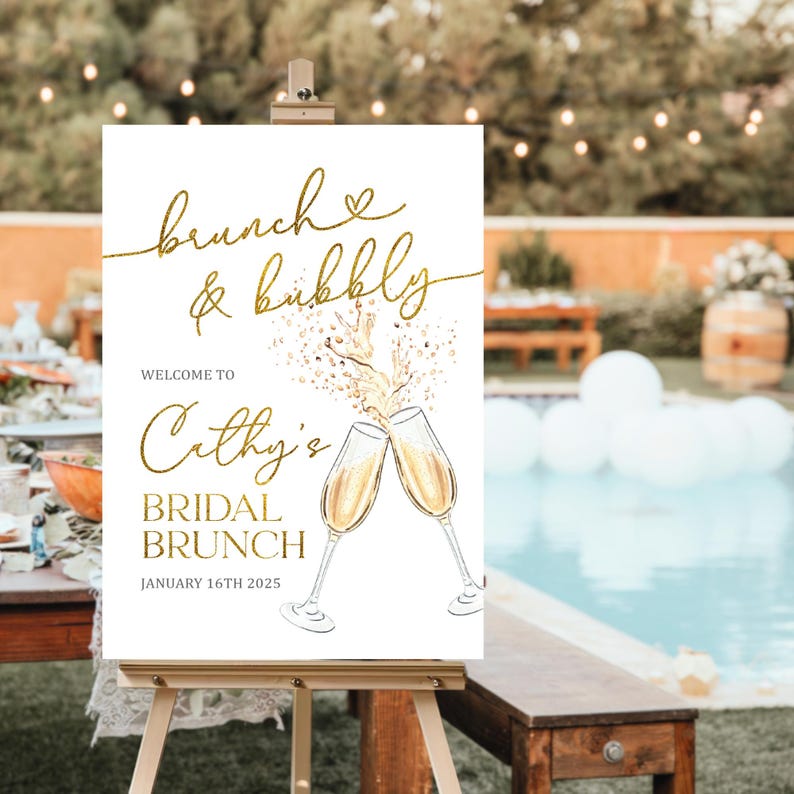 May include: A white sign with gold lettering and illustrations, displayed on a wooden easel. The sign reads "brunch & bubbly" and "Cathy's Bridal Brunch, January 16th 2025". Two champagne glasses are illustrated.