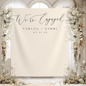 May include: A white backdrop with the text "We're Engaged" in black cursive script. Below the text, it reads "Carlos & Sammi" and "05.31.25". The backdrop is decorated with white and beige floral arrangements on either side.