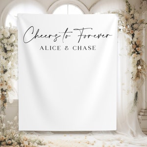 May include: White backdrop with the text "Cheers to Forever" and "Alice & Chase" in black script font. The backdrop is hanging in a room with white walls and floral decorations.