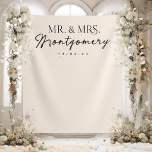 Può includere: Un fondale nuziale color crema con la scritta "MR. & MRS. Montgomery" e la data "12.03.25" in corsivo nero. Il fondale è affiancato da composizioni floreali di fiori bianchi e crema, creando un'ambientazione romantica ed elegante.