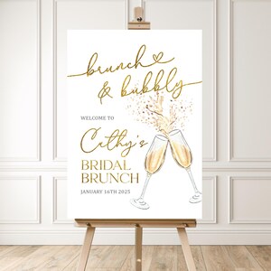 May include: A white sign on an easel with gold text that reads "brunch & bubbly" and "Cathy's Bridal Brunch". Two champagne flutes are illustrated, with champagne pouring out. The date "January 16th 2025" is also displayed.