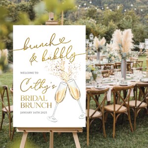 May include: A white sign on an easel with gold lettering that reads "brunch & bubbly" and "Welcome to Cathy's Bridal Brunch January 16th 2025". The sign features illustrations of champagne glasses clinking together. The background shows a brunch setting.