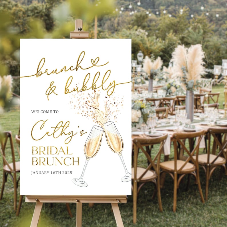 May include: A white sign on an easel with gold lettering that reads "brunch & bubbly" and "Welcome to Cathy's Bridal Brunch January 16th 2025". The sign features illustrations of champagne glasses clinking together. The background shows a brunch setting.