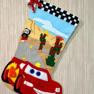May include: A handmade Christmas stocking featuring a red race car design. The stocking has a blue sky background with desert scenery, cacti, and a Route 66 sign. The top is checkered, and a red and white bow adorns the top.