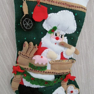 May include: A Christmas stocking featuring a felt Santa Claus chef design. The stocking is green with a white top and is decorated with sequins, a red oven mitt, and gingerbread men. The Santa Claus is holding a wooden spoon.
