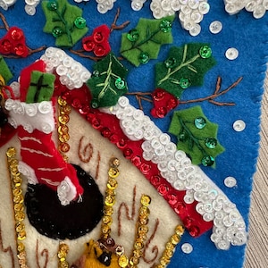 May include: A felt Christmas stocking with a blue background, white snow, green holly, red berries, and gold sequins. A birdhouse with a red roof and white trim is on the stocking. The birdhouse has a red and white candy cane hanging from it.