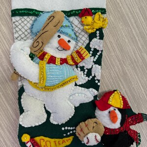 May include: Handmade Christmas stocking with a baseball theme. Features a snowman with a baseball bat, a yellow bird, and a penguin. The stocking is green with white accents and embellished with sequins.
