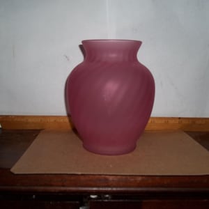 May include: A dusty rose-colored vase with a rounded body and a flared rim. The glass has a frosted appearance with subtle, swirling patterns. The vase is likely for decorative purposes or holding flowers.