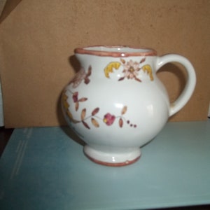 May include: A white ceramic creamer with a brown rim and floral design in shades of brown, yellow, and pink.