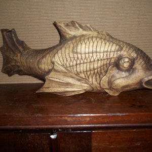 May include: A wooden carving of a fish with detailed scales and a realistic expression. The fish is painted in shades of brown and has a slightly weathered appearance.