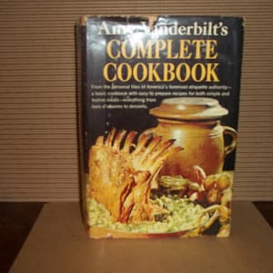 May include: A black and gold hardcover cookbook titled "Amy Vanderbilt's Complete Cookbook". The cover features a photo of a roasted leg of lamb with vegetables and a brown ceramic pot.
