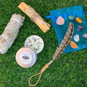 May include: A blue pouch with a variety of crystals, a white sage smudge stick, a palo santo stick, a small tin of lavender and chamomile soy candle, and a brown feather with a silver tip.
