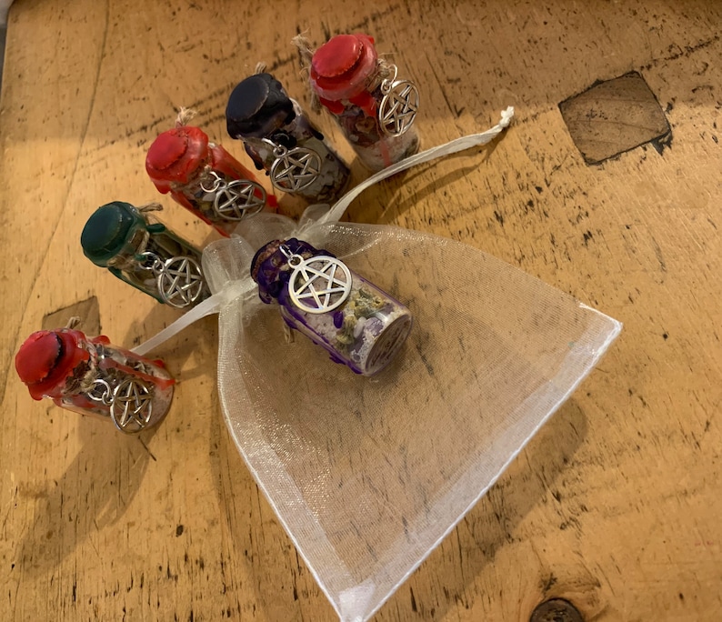 May include: Five small glass bottles filled with dried herbs and crystals, each sealed with coloured wax and featuring a silver-toned pentacle charm. One bottle is placed inside a white sheer organza drawstring pouch, while four others are arranged nearby.