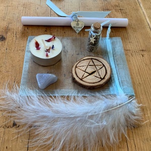 May include: A blue organza bag with a white feather, a small candle with dried flowers, a small bottle with glitter and a star charm, a wooden circle with a pentagram carved into it, a small grey stone, and a silver charm with the words "Blessed Be" on it.