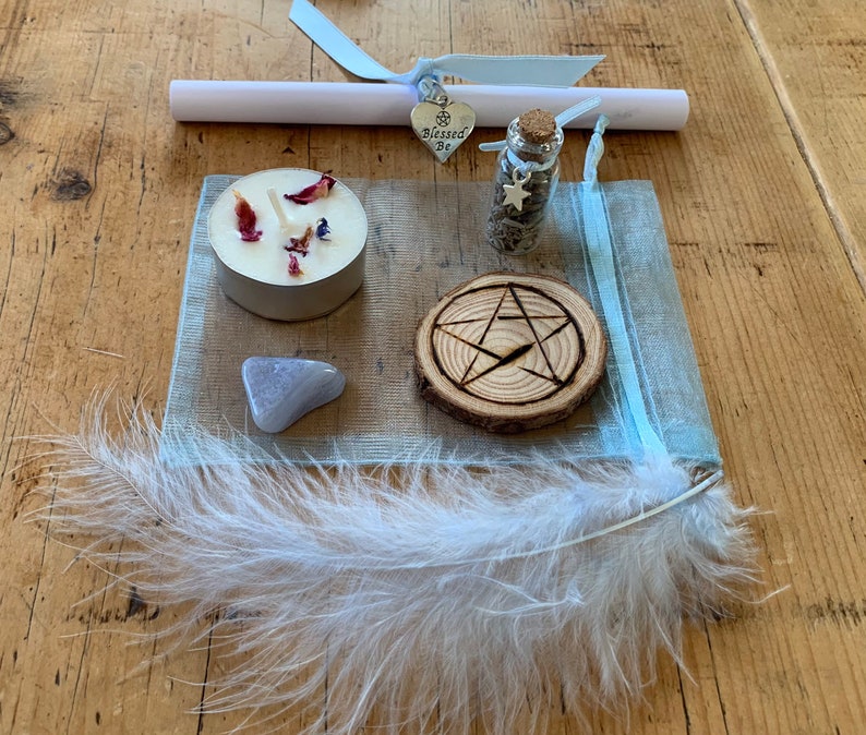 May include: A blue organza bag with a white feather, a small candle with dried flowers, a small bottle with glitter and a star charm, a wooden circle with a pentagram carved into it, a small grey stone, and a silver charm with the words "Blessed Be" on it.