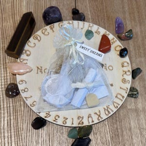 Könnte beinhalten: Ein Ouija-Brett aus Holz mit einem weißen Stoffbeutel mit der Aufschrift "SWEET DREAMS" darauf. Das Brett ist von verschiedenen farbigen Edelsteinen umgeben.