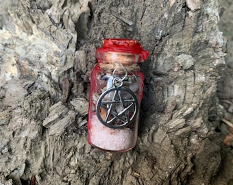 May include: A small clear glass bottle filled with pink salt and dried botanical elements, sealed with a cork and red wax. A silver-toned metal pentagram charm is attached to the neck of the bottle with a piece of natural twine wrapped around it.