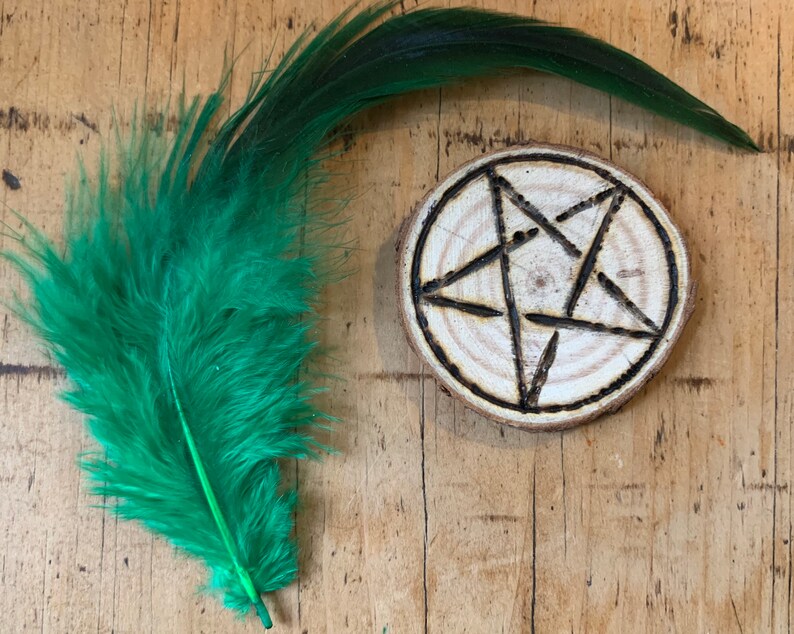 May include: A wooden circle with a five-pointed star burned into the surface. The star is a pentagram, a symbol often associated with witchcraft and paganism. A green feather is in the background.