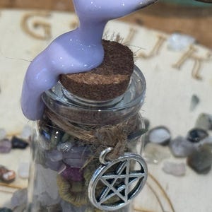 May include: A small clear glass bottle filled with assorted small colourful stones and topped with a cork stopper. A thick lavender liquid is being poured over the cork. A silver-toned metal pentagram charm is tied to the bottle neck with twine.