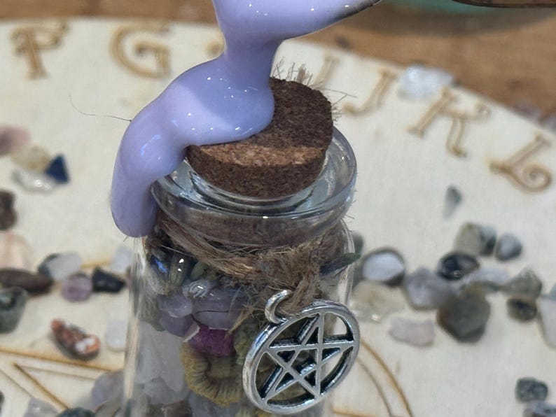 May include: A small clear glass bottle filled with assorted small colourful stones and topped with a cork stopper. A thick lavender liquid is being poured over the cork. A silver-toned metal pentagram charm is tied to the bottle neck with twine.