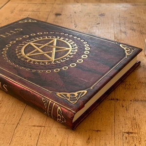 Peut inclure: Un journal reli&eacute; en cuir marron avec des accents dor&eacute;s et le mot "Spells" en relief sur la couverture. Le journal est ouvert pour r&eacute;v&eacute;ler un pentagramme avec un cercle dor&eacute; autour. Une petite bouteille en verre avec un charme d'&eacute;toile dor&eacute;e est pos&eacute;e sur un sous-verre en bois avec un motif de pentagramme. Une petite bougie avec une flamme est pos&eacute;e sur un sous-verre argent&eacute;.