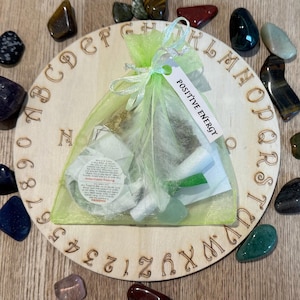Peut inclure: Un sac en organza vert avec une étiquette blanche indiquant "Positive Energy" sur un plateau en bois avec des lettres et des chiffres autour du bord. Le sac contient divers cristaux et pierres.