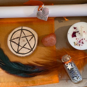 May include: A small orange box with a variety of items inside, including a wooden circle with a pentagram, a small orange stone, a white candle with dried rose petals, a small glass bottle with lavender and a star, a feather, and a white scroll with a silver heart charm that says "Blessed Be".