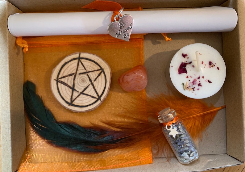 May include: A small orange box with a variety of items inside, including a wooden circle with a pentagram, a small orange stone, a white candle with dried rose petals, a small glass bottle with lavender and a star, a feather, and a white scroll with a silver heart charm that says "Blessed Be".