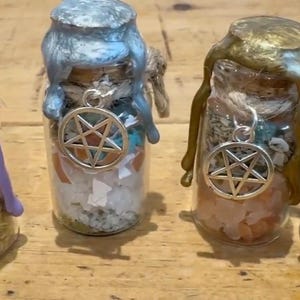 May include: Four small glass jars filled with various crystals, herbs, and salt, each sealed with a cork and coloured wax. A silver-toned pentagram charm hangs from a twine string tied around the neck of each bottle. The wax colours are purple, light blue, gold, and grey.