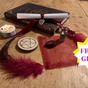 May include: A witchy spell kit with a book of spells, a rolled up scroll tied with a burgundy ribbon, a small candle, a wooden circle with a pentagram, a red feather, a small red pouch, a small glass bottle with a silver star charm, and a red heart-shaped stone. The kit is on a wooden surface. The text "FREE GIFT" is in a yellow starburst.