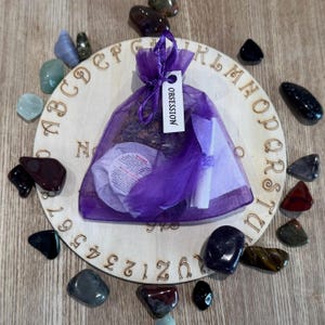 May include: A wooden Ouija board with a purple pouch labeled "Obsession" and various colorful stones surrounding it.