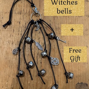 May include: A hanging ornament featuring multiple black cords attached to a silver metal ring and lobster clasp. The cords are adorned with silver-toned charms including a crescent moon, a feather, a key, a pentagram, and a Celtic knot, plus several small round bells. Text reads Witches bells + Free Gift.