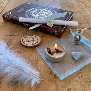 May include: A witchy spell kit with a book titled "Spells", a white feather, a wooden coaster with a pentagram, a small glass bottle with herbs, a tealight candle, a blue organza bag, and a silver charm with the words "Be Blessed" on it.