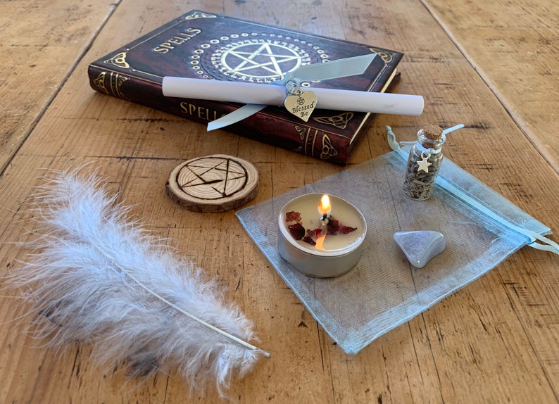 May include: A witchy spell kit with a book titled "Spells", a white feather, a wooden coaster with a pentagram, a small glass bottle with herbs, a tealight candle, a blue organza bag, and a silver charm with the words "Be Blessed" on it.