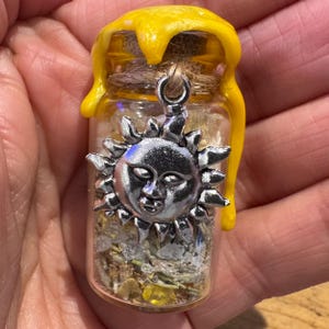 May include: A small glass jar filled with dried herbs and crystals, topped with a yellow wax seal that drips down the sides. A silver-toned metal sun charm with a face is attached to the neck of the jar with a piece of brown twine.
