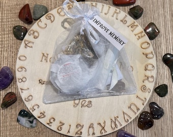 May include: A wooden circular board with letters and numbers around the edge. A white organza bag with a white label that says "IMPROVE MEMORY" is on top of the board. The bag contains a small white pouch and a brown herbal tea blend. The board is surrounded by various colored stones.
