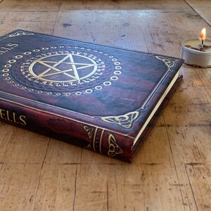 May include: A brown leather-bound book with gold lettering that reads "Spells" on the cover. The book is open and shows a gold pentagram with a circle of runes around it. There is a small glass bottle with a cork stopper and a silver star charm on a wooden base with a pentagram carved into it. A small white candle is lit in the background.