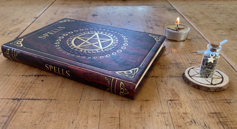 May include: A brown leather-bound book with gold lettering that reads "Spells" on the cover. The book is open and shows a gold pentagram with a circle of runes around it. There is a small glass bottle with a cork stopper and a silver star charm on a wooden base with a pentagram carved into it. A small white candle is lit in the background.