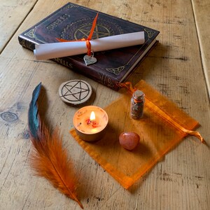 May include: A witchy spell kit with a brown leather-bound book with the word "Spells" on the cover, a white scroll, a small heart charm, a wooden pentagram, a small glass bottle with cork stopper, a small orange candle, a small orange crystal, and a brown and orange feather.