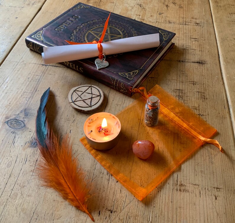 May include: A witchy spell kit with a brown leather-bound book with the word "Spells" on the cover, a white scroll, a small heart charm, a wooden pentagram, a small glass bottle with cork stopper, a small orange candle, a small orange crystal, and a brown and orange feather.