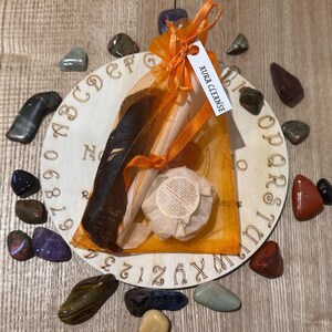 May include: A wooden circular board with letters and numbers around the edge. A small orange pouch with a white tag that says "Aura Cleanse" is on top of the board. The pouch contains a feather and a small white container. There are several colorful stones around the board.