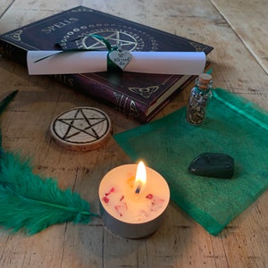May include: A green feather, a small book with the word "Spells" on the cover, a wooden pentagram, a small bottle with a cork stopper, a green pouch, a green heart charm with the words "Blessed Be" on it, a white candle with red flakes, and a dark green stone.