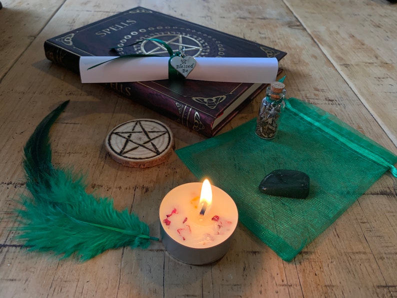 May include: A green feather, a small book with the word "Spells" on the cover, a wooden pentagram, a small bottle with a cork stopper, a green pouch, a green heart charm with the words "Blessed Be" on it, a white candle with red flakes, and a dark green stone.