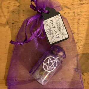 May include: A purple organza drawstring pouch containing a small glass bottle with a cork stopper. The bottle features a silver-toned pentagram charm attached to the exterior. A black paper tag tied to the pouch is inscribed with the word ANXIETY.