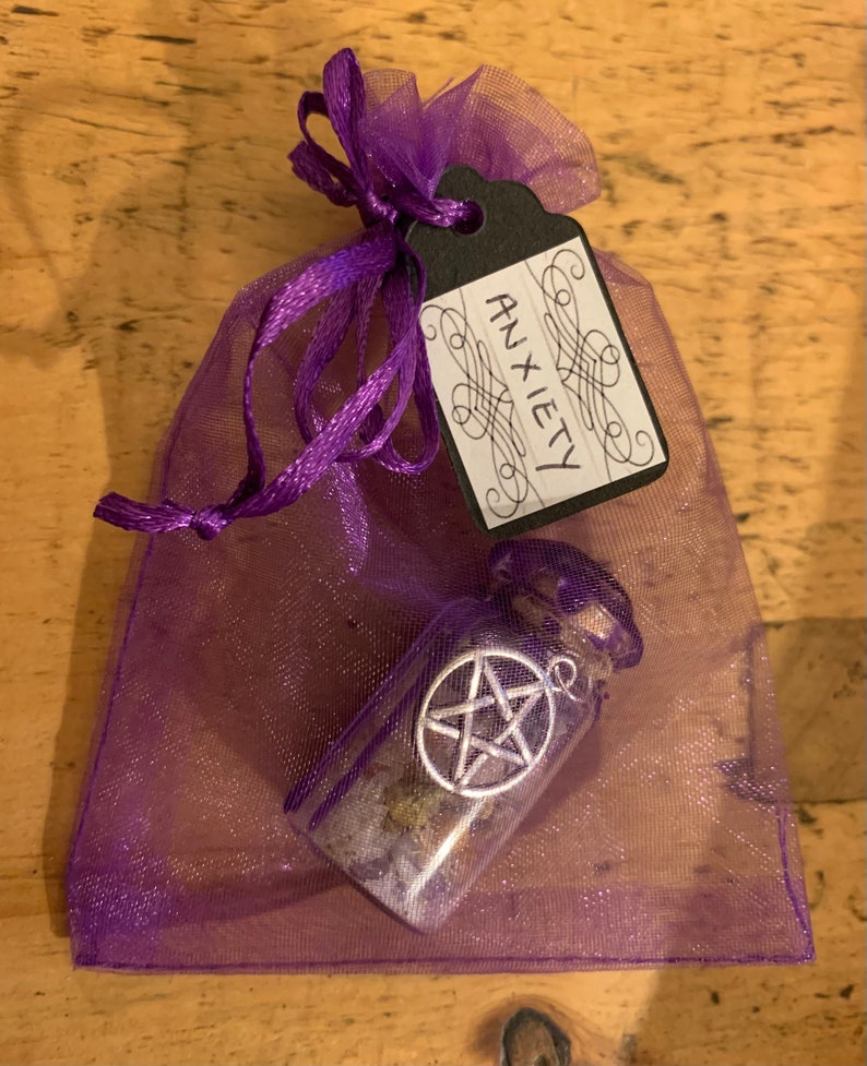 May include: A purple organza drawstring pouch containing a small glass bottle with a cork stopper. The bottle features a silver-toned pentagram charm attached to the exterior. A black paper tag tied to the pouch is inscribed with the word ANXIETY.