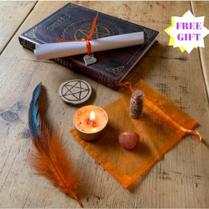 May include: A witchy spell kit with a brown leather-bound book with the word "Spells" on the cover, a white scroll tied with an orange ribbon, a small orange feather, a wooden pentagram, a small orange candle, a small orange pouch with a glass vial of herbs, and a red stone.