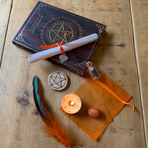 May include: A witchy spell book with a pentagram on the cover, a rolled up scroll, a small wooden pentagram, a small glass bottle with glitter, a small orange candle, an orange stone, and a feather with blue and orange colors.
