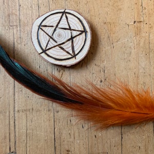 May include: A wooden circle with a pentagram burned into the surface, and a long orange and black feather lying on a wooden surface.