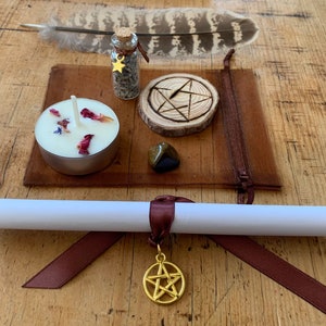 Peut inclure: Une pochette en cuir marron contenant une bougie blanche, une petite bouteille en verre avec un charme d'&eacute;toile dor&eacute;e, un cercle en bois avec un pentagramme grav&eacute;, une plume brune et une pierre brun fonc&eacute;. La pochette est sur une surface en bois.